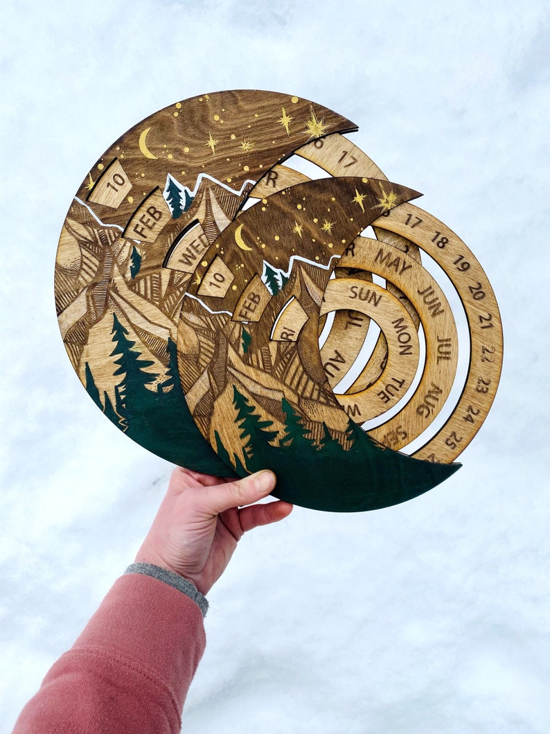 Peut inclure: Calendrier en bois avec un motif de montagne et de for&ecirc;t. Le calendrier a la forme d'un croissant de lune et poss&egrave;de un cadran rotatif pour chaque mois. Le calendrier est en bois et a une palette de couleurs verte et brune.