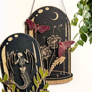 May include: Two wooden wall hangings with a black background and a glass dome design. The first features a skeleton mermaid with green leaves. The second features a moth and dandelion with a crescent moon in the background.