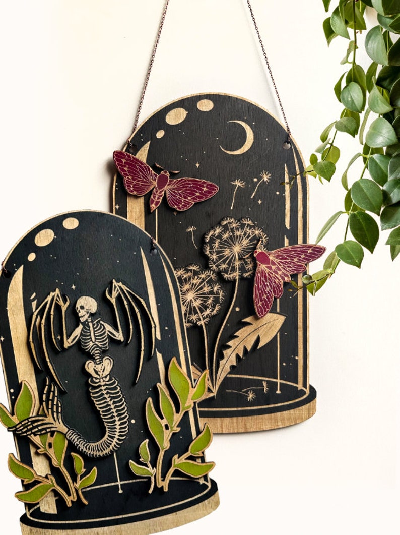 May include: Two wooden wall hangings with a black background and a glass dome design. The first features a skeleton mermaid with green leaves. The second features a moth and dandelion with a crescent moon in the background.