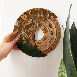Peut inclure: Un calendrier en bois avec un motif de montagne et de for&ecirc;t. Le calendrier est en forme de cercle et a une lune croissante et des &eacute;toiles en haut. Le calendrier est divis&eacute; en mois, avec les jours de la semaine list&eacute;s sur l'anneau ext&eacute;rieur. Les mois sont janvier, f&eacute;vrier, mars, avril, mai, juin, juillet, ao&ucirc;t, septembre, octobre, novembre et d&eacute;cembre.