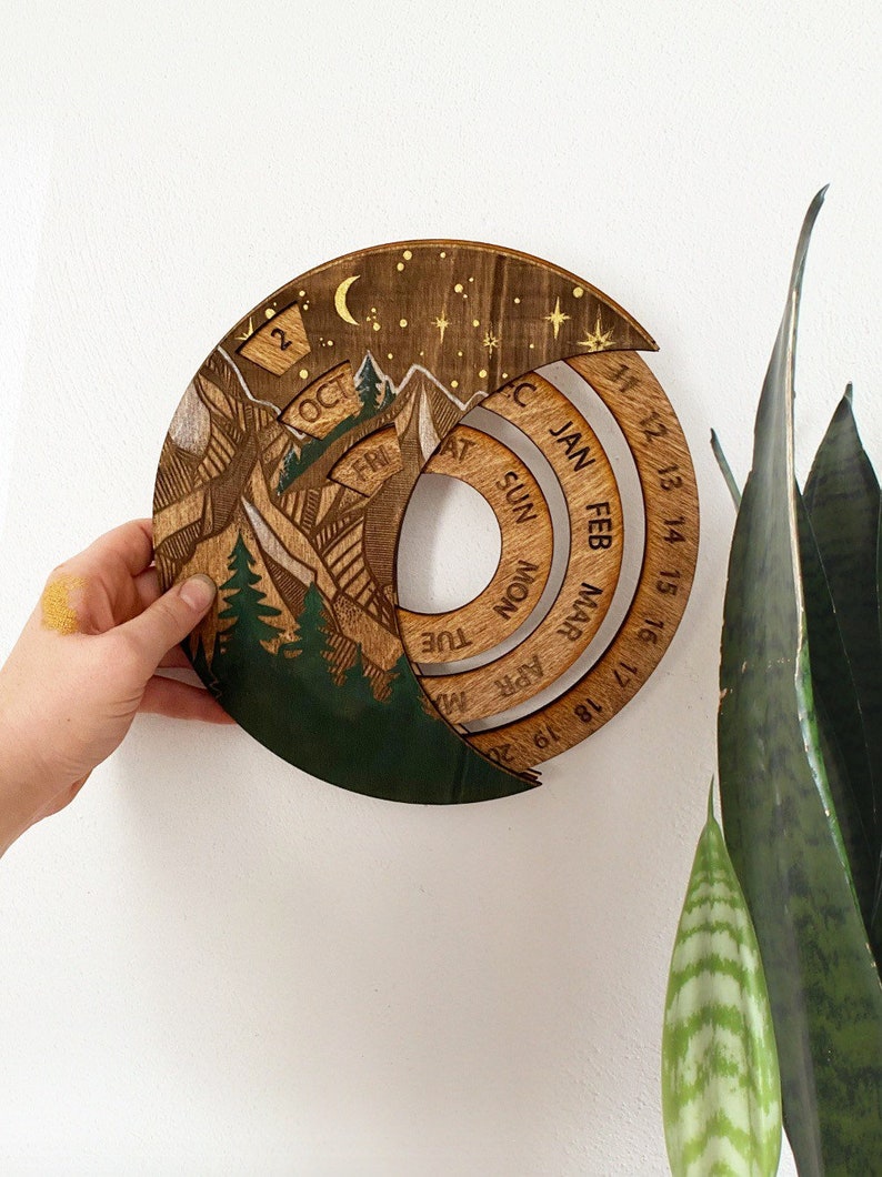 Peut inclure: Un calendrier en bois avec un motif de montagne et de for&ecirc;t. Le calendrier est en forme de cercle et a une lune croissante et des &eacute;toiles en haut. Le calendrier est divis&eacute; en mois, avec les jours de la semaine list&eacute;s sur l'anneau ext&eacute;rieur. Les mois sont janvier, f&eacute;vrier, mars, avril, mai, juin, juillet, ao&ucirc;t, septembre, octobre, novembre et d&eacute;cembre.