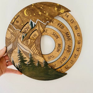 Peut inclure: Un calendrier en bois avec une sc&egrave;ne de montagne et un ciel nocturne &eacute;toil&eacute;. Le calendrier pr&eacute;sente un design circulaire avec des anneaux rotatifs pour chaque mois de l'ann&eacute;e. Les mois sont &eacute;tiquet&eacute;s en cercle, en commen&ccedil;ant par f&eacute;vrier et en terminant par janvier. Le calendrier est en bois et a une finition naturelle.