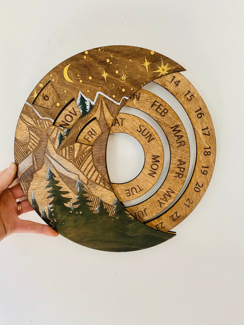 Peut inclure: Un calendrier en bois avec une sc&egrave;ne de montagne et un ciel nocturne &eacute;toil&eacute;. Le calendrier pr&eacute;sente un design circulaire avec des anneaux rotatifs pour chaque mois de l'ann&eacute;e. Les mois sont &eacute;tiquet&eacute;s en cercle, en commen&ccedil;ant par f&eacute;vrier et en terminant par janvier. Le calendrier est en bois et a une finition naturelle.
