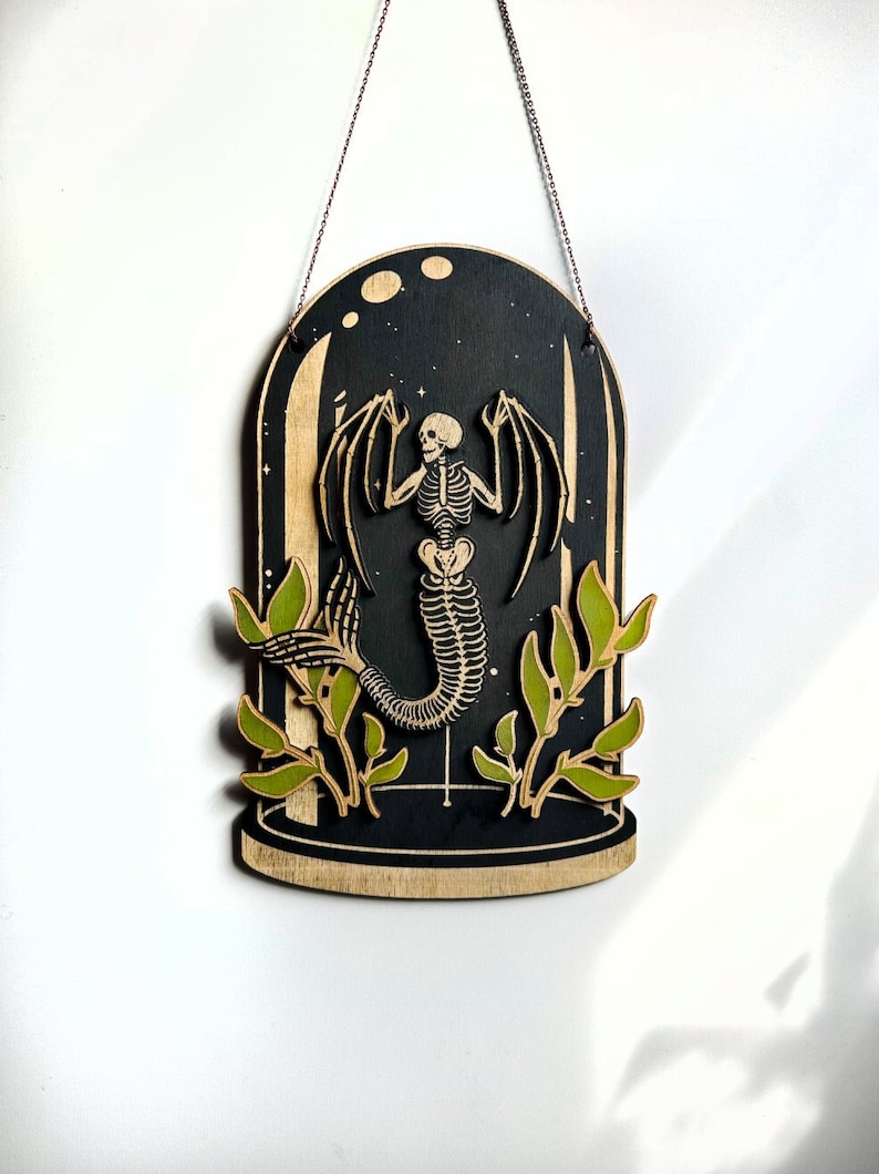 May include: A wooden wall hanging featuring a skeletal mermaid with wings inside a glass dome. The mermaid is surrounded by green leaves and the background is black with white stars.