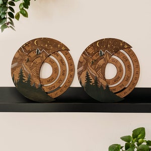 Peut inclure: Deux calendriers perp&eacute;tuels en bois avec un motif de montagne et de for&ecirc;t. Les calendriers sont circulaires et pr&eacute;sentent les mois de l'ann&eacute;e en spirale. Les calendriers sont sur une &eacute;tag&egrave;re noire.