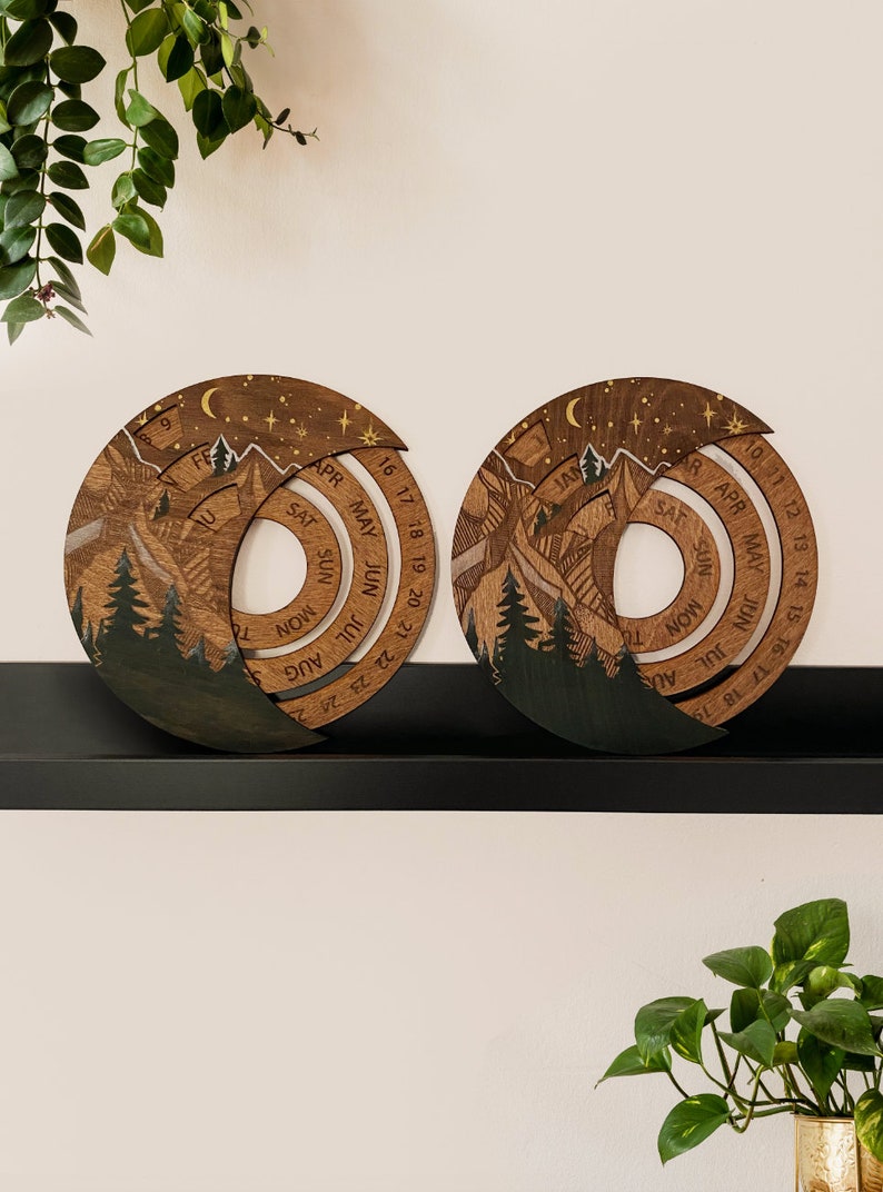 Peut inclure: Deux calendriers perp&eacute;tuels en bois avec un motif de montagne et de for&ecirc;t. Les calendriers sont circulaires et pr&eacute;sentent les mois de l'ann&eacute;e en spirale. Les calendriers sont sur une &eacute;tag&egrave;re noire.