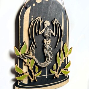 May include: A wooden wall hanging featuring a skeletal mermaid with wings inside a glass dome. The mermaid is surrounded by green leaves.