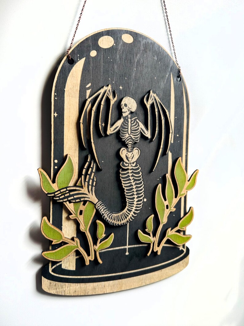 May include: A wooden wall hanging featuring a skeletal mermaid with wings inside a glass dome. The mermaid is surrounded by green leaves.