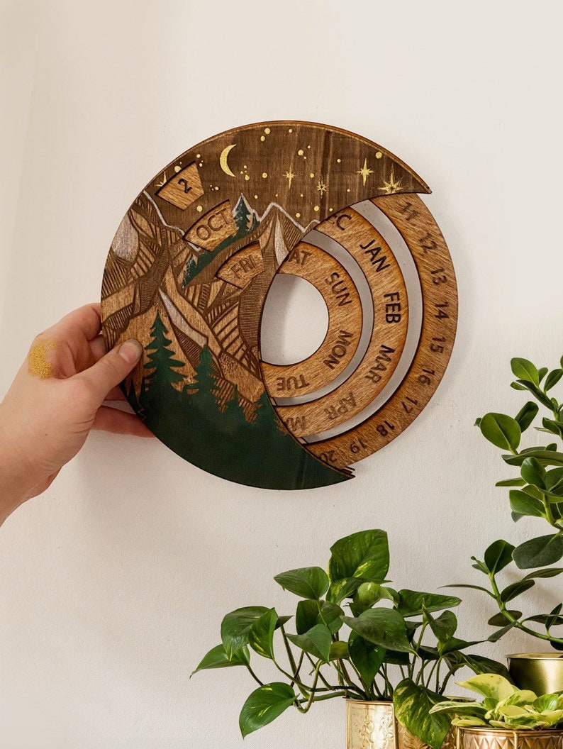 Peut inclure: Un calendrier en bois avec une sc&egrave;ne de montagne et une lune croissante. Le calendrier est en forme de cercle et a un anneau au centre. Le calendrier est divis&eacute; en mois et en jours. Les mois sont &eacute;tiquet&eacute;s avec les trois premi&egrave;res lettres du mois, et les jours sont num&eacute;rot&eacute;s de 11 &agrave; 20. Le calendrier est en bois et a une teinte brun fonc&eacute;.