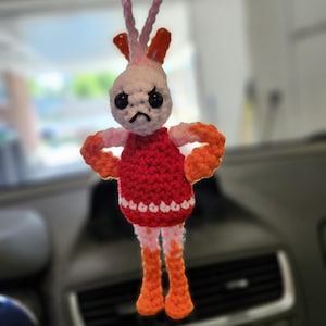 May include: A crocheted car charm depicting an angry character with a red dress, orange limbs, and white details. The charm is suspended by a pink and white cord, suitable for hanging from a car's rearview mirror. The charm is approximately 10 cm tall.