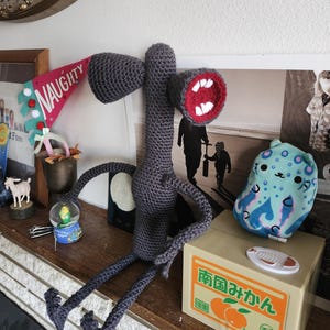 Op de afbeelding: Een handgemaakte, grijze Siren Head-knuffel met grote, luidsprekerachtige hoofden en een rood-witte mond. De knuffel zit op een houten plank naast een doos met Japanse tekst en een blauwe octopus-knuffel.