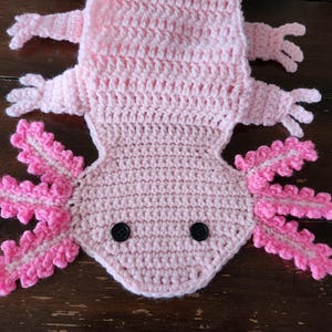 Peut inclure: Un axolotl en crochet rose avec des yeux en boutons noirs. L'axolotl a des branchies roses frisées et est crocheté en point texturé.
