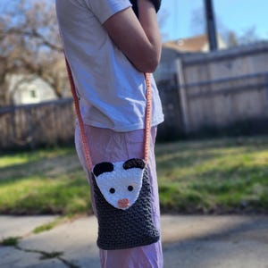 May include: A handmade crochet crossbody bag with a possum design. The bag has a grey body, a white face with black ears, and a pink nose. The strap is a light orange colour. The bag is worn over the shoulder.