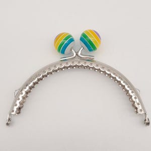 May include: A silver-toned purse frame with a curved design and decorative edge. Two colorful, spherical accents with rainbow stripes sit atop the frame. The frame has small loops on each end for attachment.