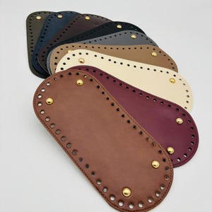 May include: A collection of oval-shaped bag bases in various colors, including brown, burgundy, cream, and black. Each base has pre-drilled holes for stitching and is adorned with gold-colored studs. These bases are designed for crafting handbags and purses.