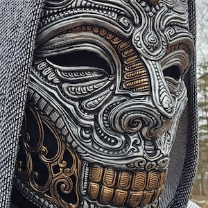 May include: A silver and gold skull mask with intricate details. The mask has a hooded design and is made of a hard material.