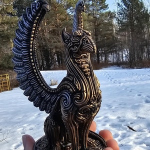 May include: A detailed, intricately designed, gold and black metal sculpture of a mythical creature with wings. The sculpture is held in a person's hand against a snowy background.