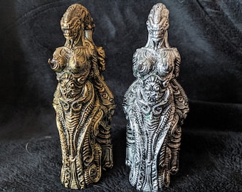 May include: Two detailed, intricately sculpted figurines of a woman with a futuristic, alien-like design. One is gold, the other is silver.