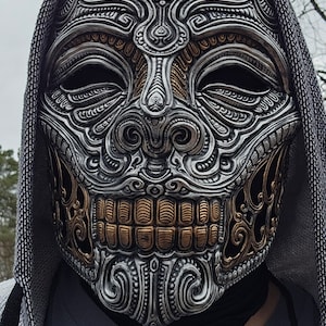 Op de afbeelding: Een zilveren en gouden schedelmasker met ingewikkelde details en een capuchon.