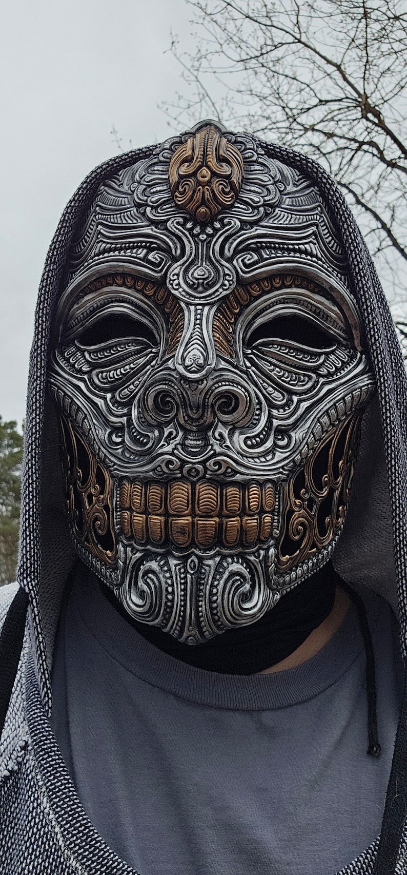 Op de afbeelding: Een zilveren en gouden schedelmasker met ingewikkelde details en een capuchon.