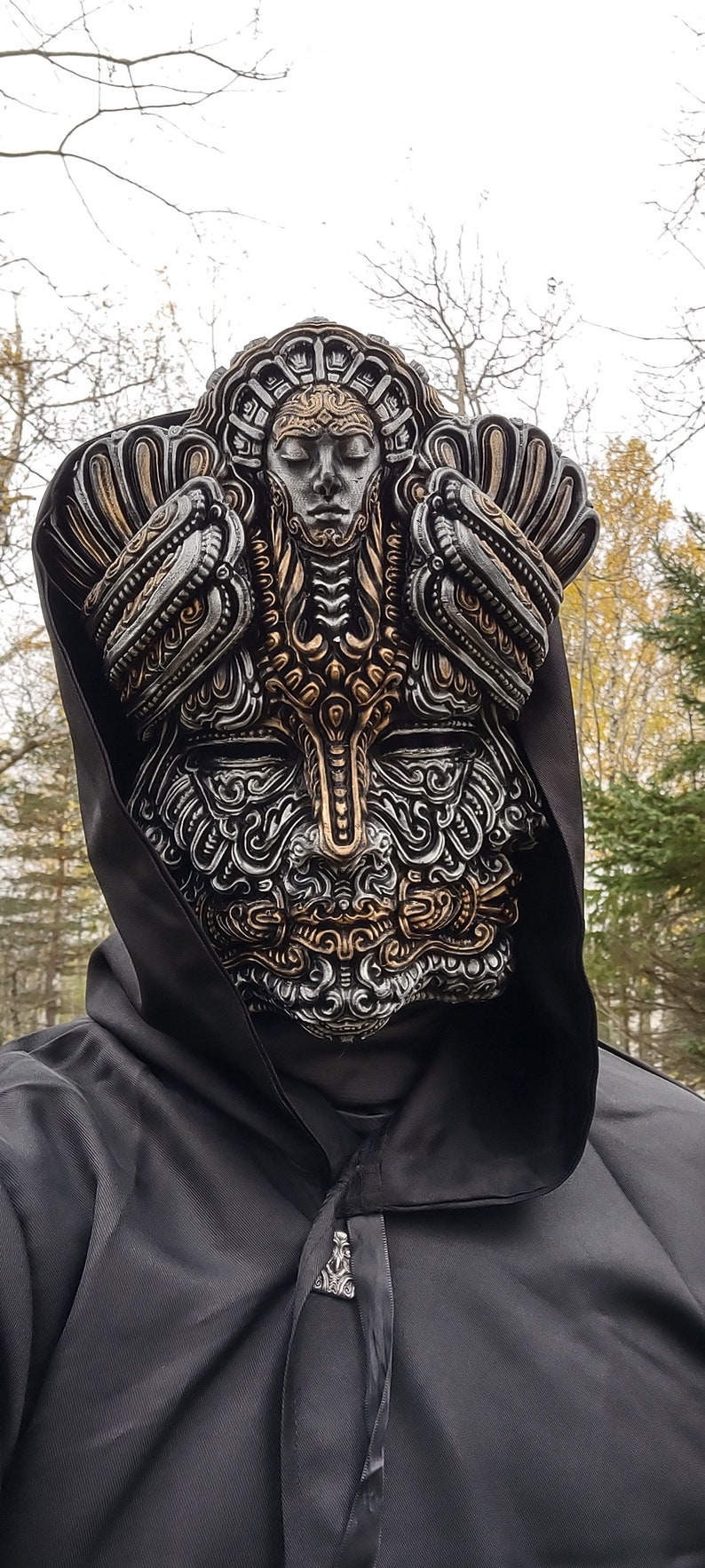 May include: A person wearing a black hooded cloak with a silver and gold ornate mask covering their face. The mask features intricate details and a human-like face.