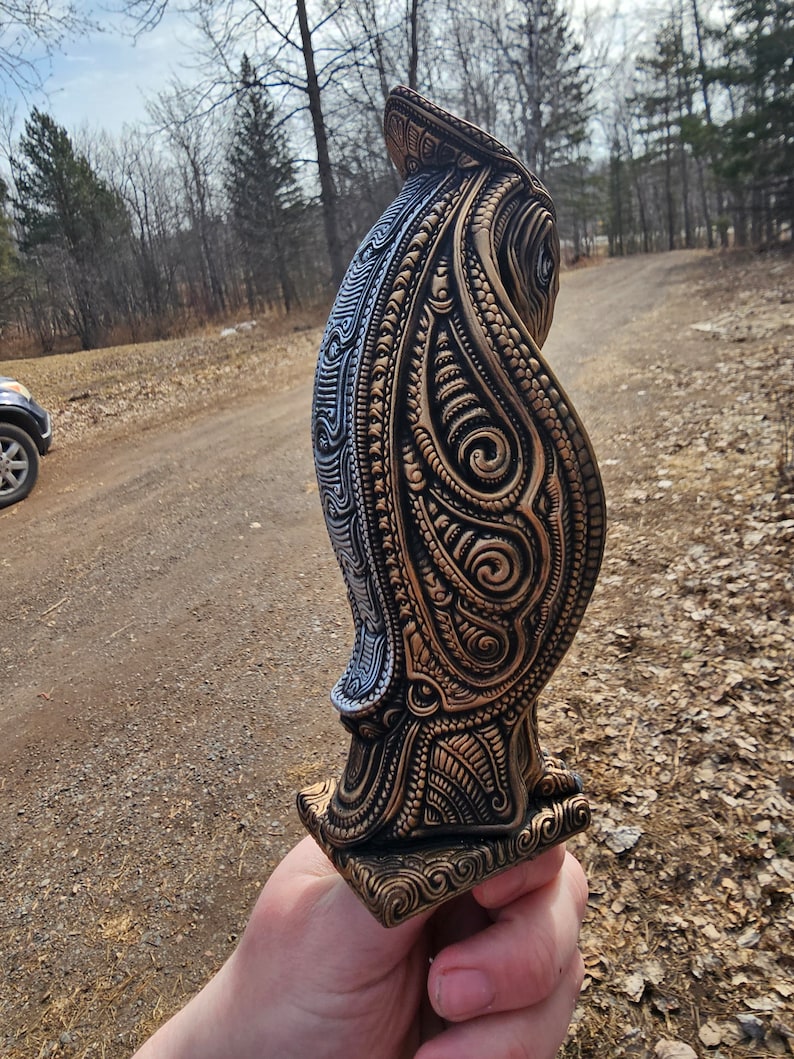 May include: A detailed, intricately carved wooden owl figurine with gold and silver paint. The owl is standing on a square base and has a detailed design on its feathers.