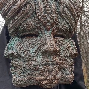 May include: A detailed, ornate, bronze mask with a green patina. The mask features a human face with intricate carvings and a smaller face within the forehead.