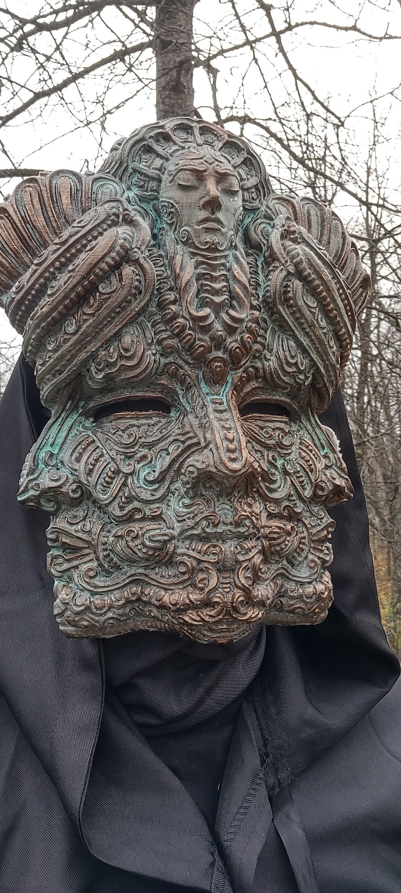 May include: A detailed, ornate, bronze mask with a green patina. The mask features a human face with intricate carvings and a smaller face within the forehead.