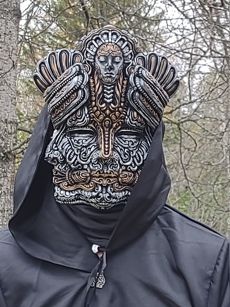 May include: A silver and gold detailed mask with intricate designs. The mask features a human face with closed eyes and a crown-like top.