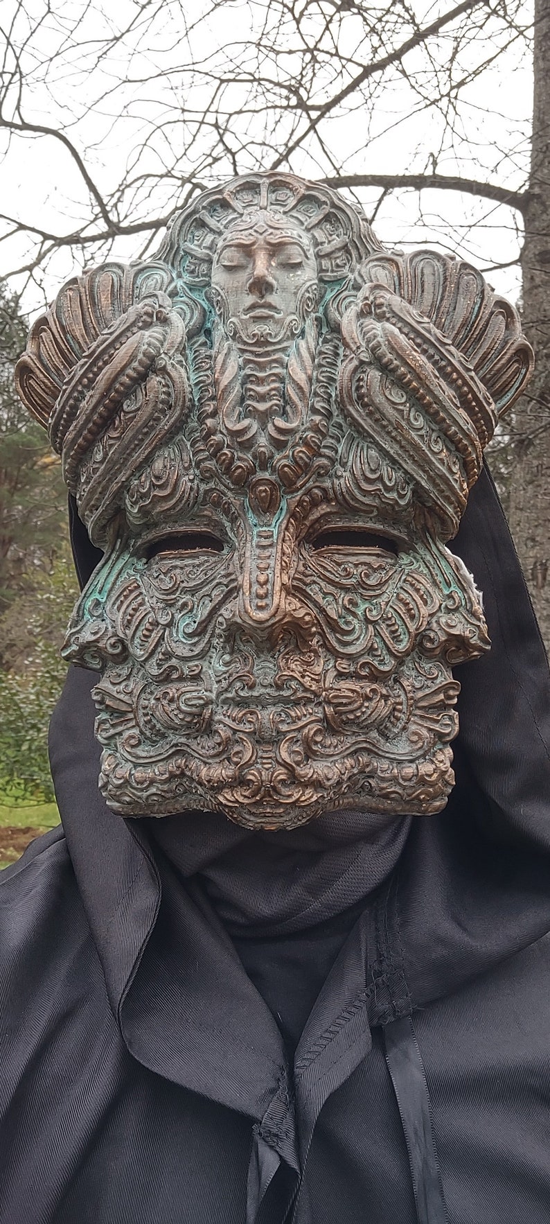 May include: A detailed, ornate mask with a copper and green patina. The mask features a human face with intricate carvings and a crown-like top.