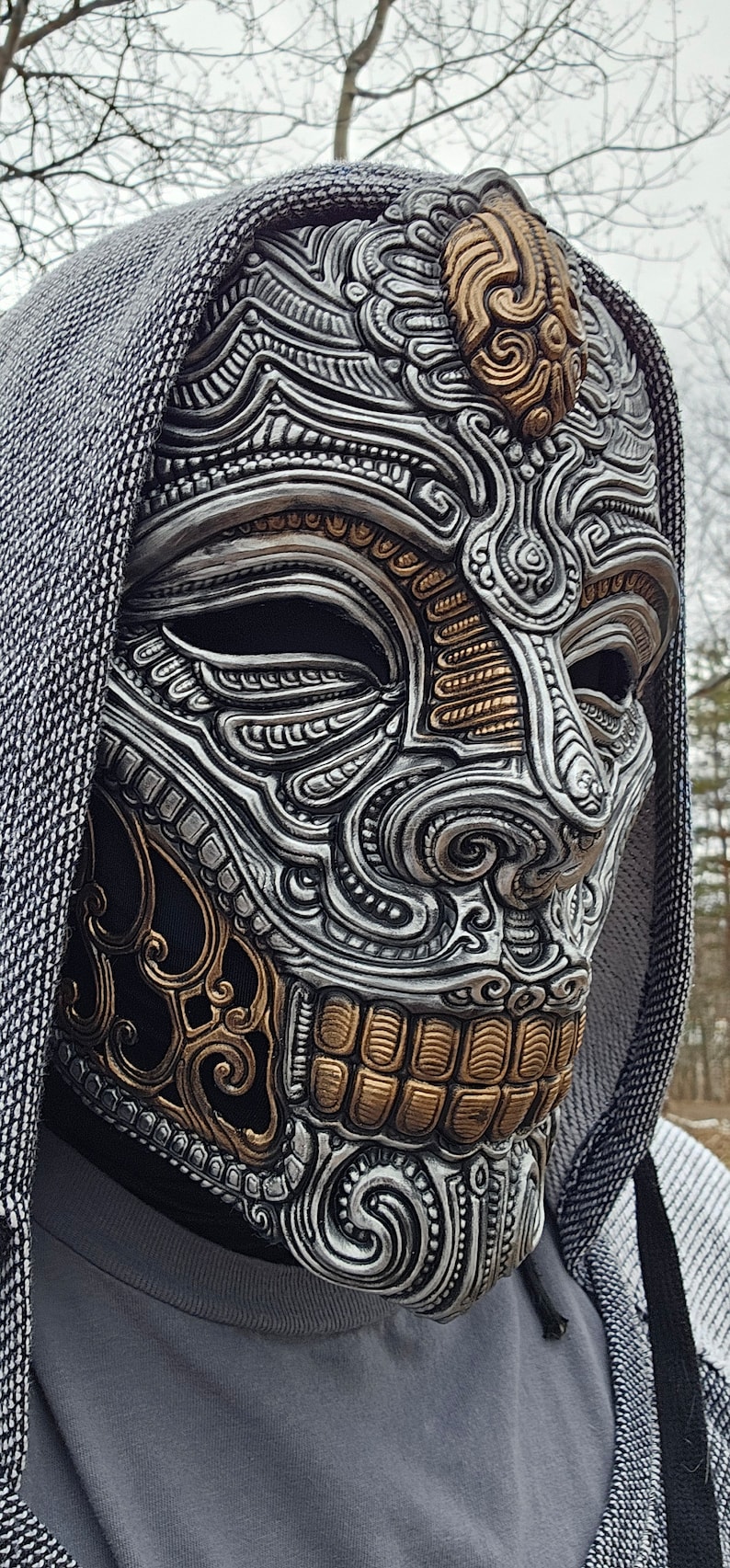 May include: A silver and gold skull mask with intricate details. The mask has a hooded design and is made of a hard material.