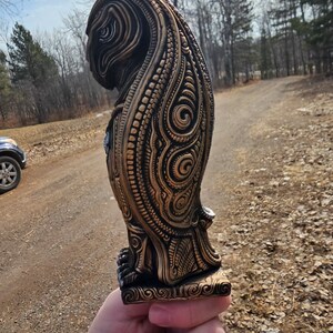 May include: A detailed, intricately carved owl statue with a gold finish. The owl is standing on a base with a decorative design. The statue is made of a dark material and has a textured surface.