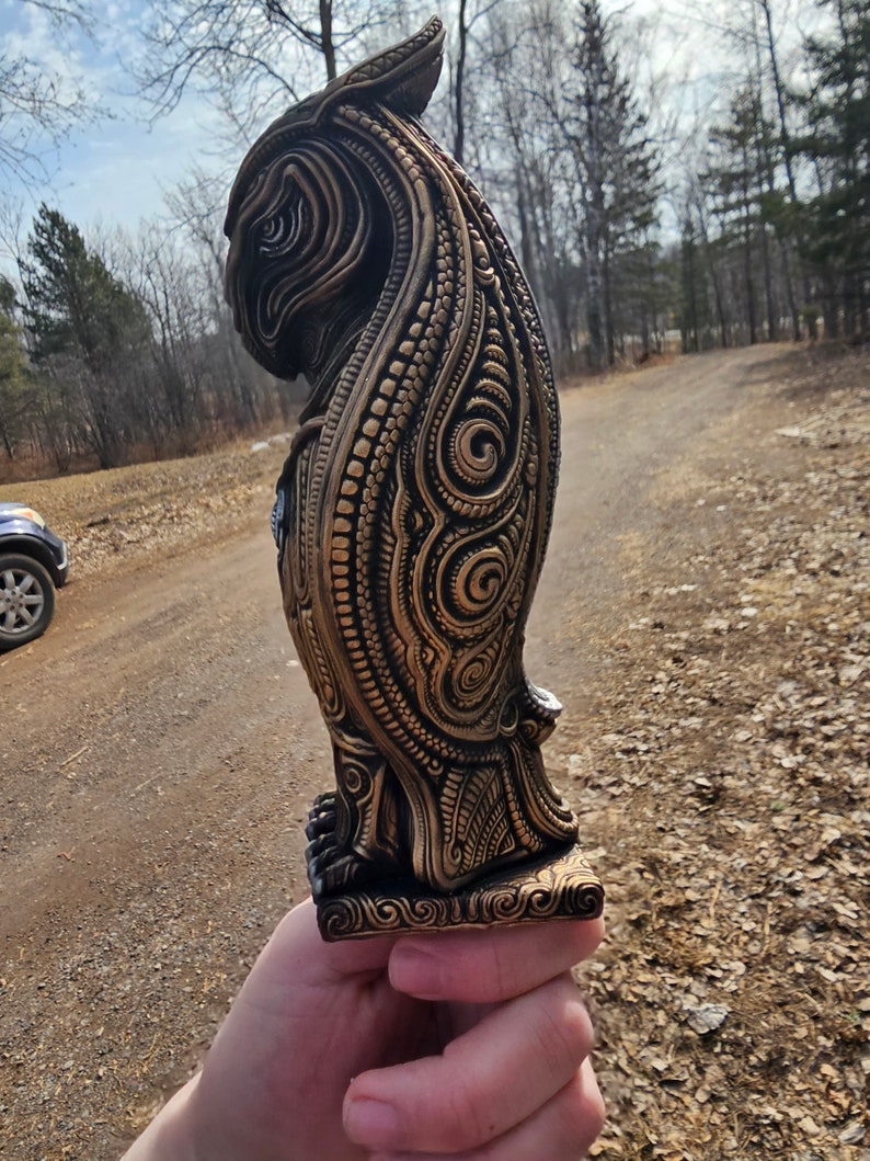 May include: A detailed, intricately carved owl statue with a gold finish. The owl is standing on a base with a decorative design. The statue is made of a dark material and has a textured surface.