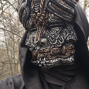 May include: A person wearing a black hooded cloak with a detailed, silver and gold mask covering their face. The mask features intricate designs and a human-like face with closed eyes.