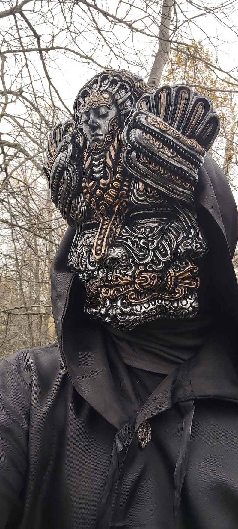 May include: A person wearing a black hooded cloak with a detailed, silver and gold mask covering their face. The mask features intricate designs and a human-like face with closed eyes.