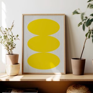 May include: A framed print with a minimalist design featuring three stacked yellow ovals on a white background. The print is displayed on a wooden shelf with potted plants and books.