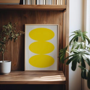 May include: A framed print with a minimalist design featuring three stacked yellow ovals on a white background.