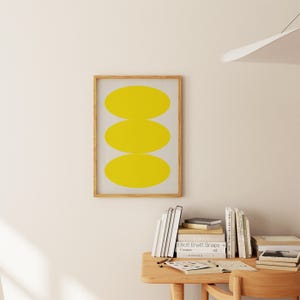 May include: A framed print with a minimalist design featuring three stacked yellow ovals on a white background.