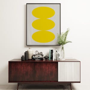 May include: A framed print with three yellow circles stacked on top of each other on a gray background. The print is sitting on a dark wood and white cabinet with books and decorative objects.