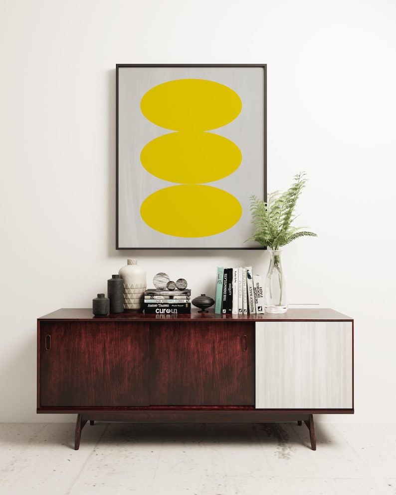 May include: A framed print with three yellow circles stacked on top of each other on a gray background. The print is sitting on a dark wood and white cabinet with books and decorative objects.