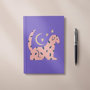 Peut inclure: Un carnet violet avec un motif de créature rose, un croissant de lune jaune et des étoiles. Un stylo noir est placé à droite. Le carnet a une couverture rigide et des pages vierges.