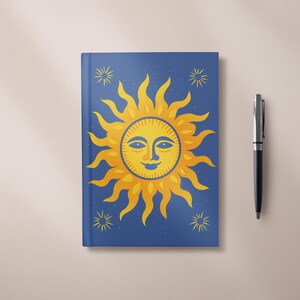 Peut inclure: Un journal bleu avec un motif de soleil jaune et des étoiles. Le soleil a un visage souriant. Un stylo argenté et noir est à côté du journal.