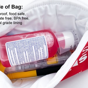 Op de afbeelding: Een rode ritszak met het woord "55" erop gedrukt. De zak is open en bevat een roze fles met een witte dop, een gele marker en andere items. De zak is gelabeld met "Inside of Bag: Waterproof, food safe, phthalate free, BPA free, medical grade lining."
