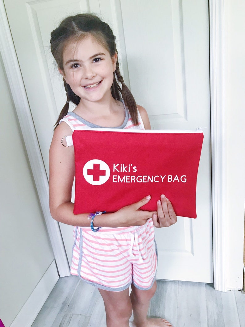 Puede incluir: Una ni&ntilde;a sostiene una bolsa roja con cremallera, con una cruz blanca y el texto "Kiki's EMERGENCY BAG".