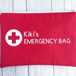 Puede incluir: Bolsa roja con cremallera con una cruz blanca en un c&iacute;rculo y el texto "Kiki's EMERGENCY BAG" en blanco.