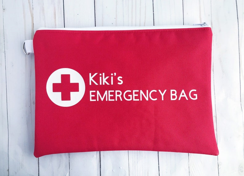 Puede incluir: Bolsa roja con cremallera con una cruz blanca en un c&iacute;rculo y el texto "Kiki's EMERGENCY BAG" en blanco.