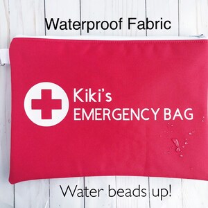Puede incluir: Bolsa roja con cremallera con una cruz blanca y el texto "Kiki's EMERGENCY BAG" impreso en ella. La bolsa est&aacute; hecha de tela impermeable y tiene gotas de agua.