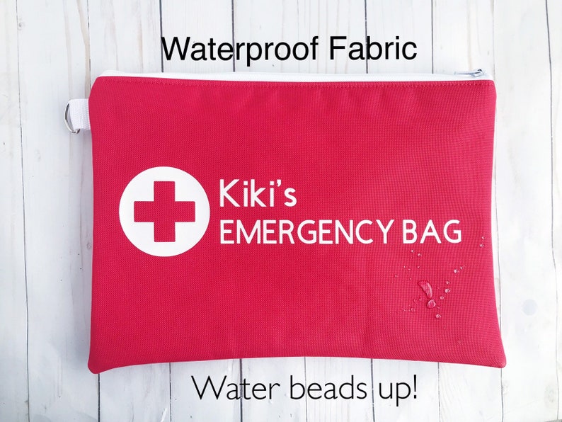 Puede incluir: Bolsa roja con cremallera con una cruz blanca y el texto "Kiki's EMERGENCY BAG" impreso en ella. La bolsa est&aacute; hecha de tela impermeable y tiene gotas de agua.