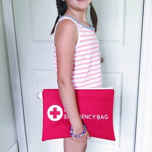 Puede incluir: Una persona joven sosteniendo una bolsa roja con cremallera con una cruz blanca y el texto "Emergency Bag".