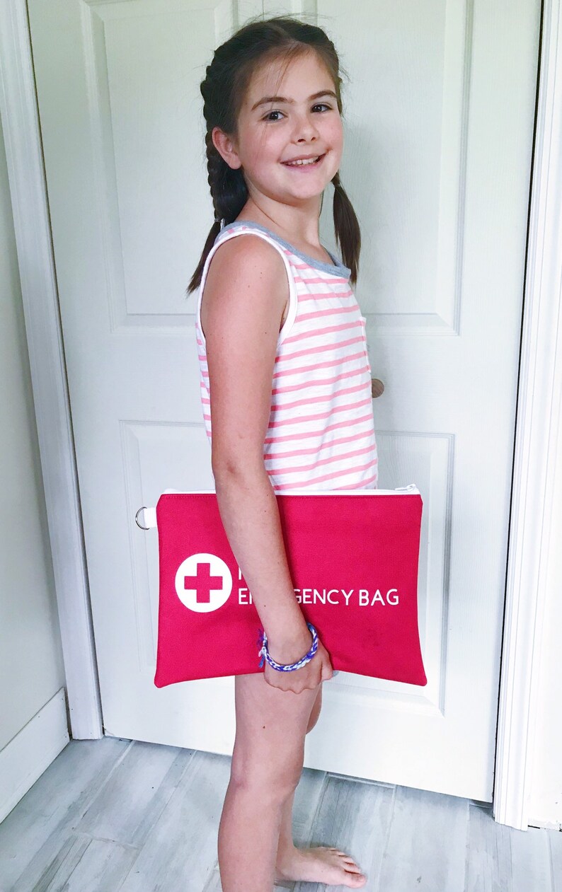 Puede incluir: Una persona joven sosteniendo una bolsa roja con cremallera con una cruz blanca y el texto "Emergency Bag".